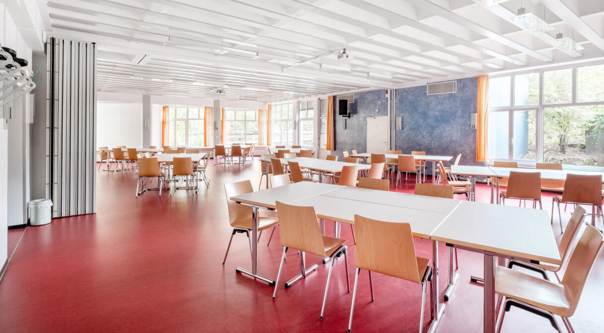 Heller Essensaal mit rotem Linoleumboden, Tischen und St&uuml;hlen. Gro&szlig;e Fensterfront.