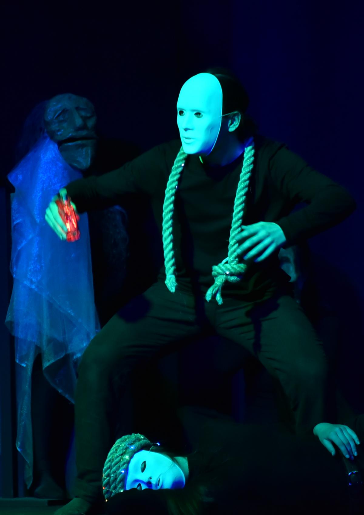 Titelbild: Eine schwarz gekleidete Figur mit einer weißen Maske und einem Tau um den Hals und einer roten Flasche steht herausfordernd im grünen Licht. Im Hintergrund weitere maskierte Figuren.