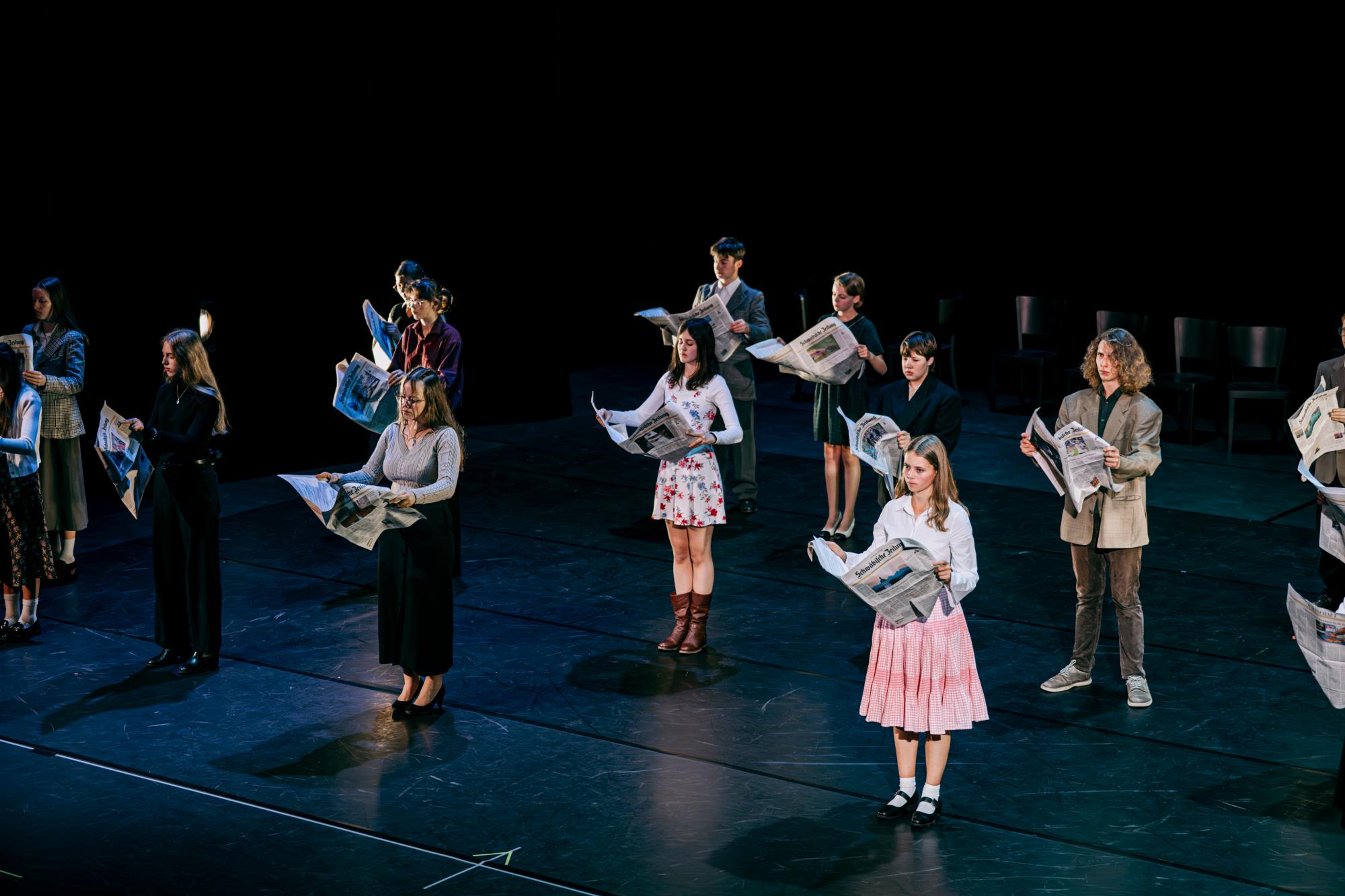 Das Ensemble steht verteilt auf der Bühne. Jede Person hat eine Zeitung aufgeschlagen vor sich.