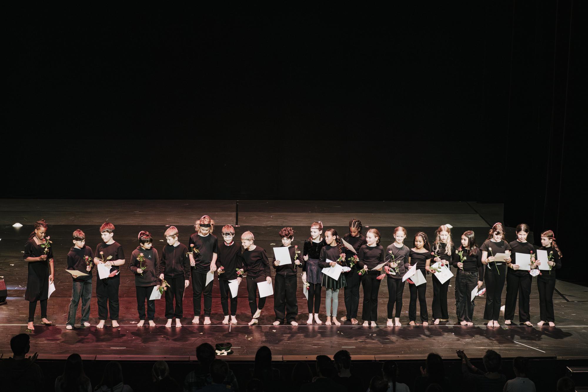 Die Grundschüler:innen stehen in einer Reihe am Bühnenrand und halten in ihrer Hand jeweils eine Urkunde und eine Blume. Teilweise gucken sie sich an. 
