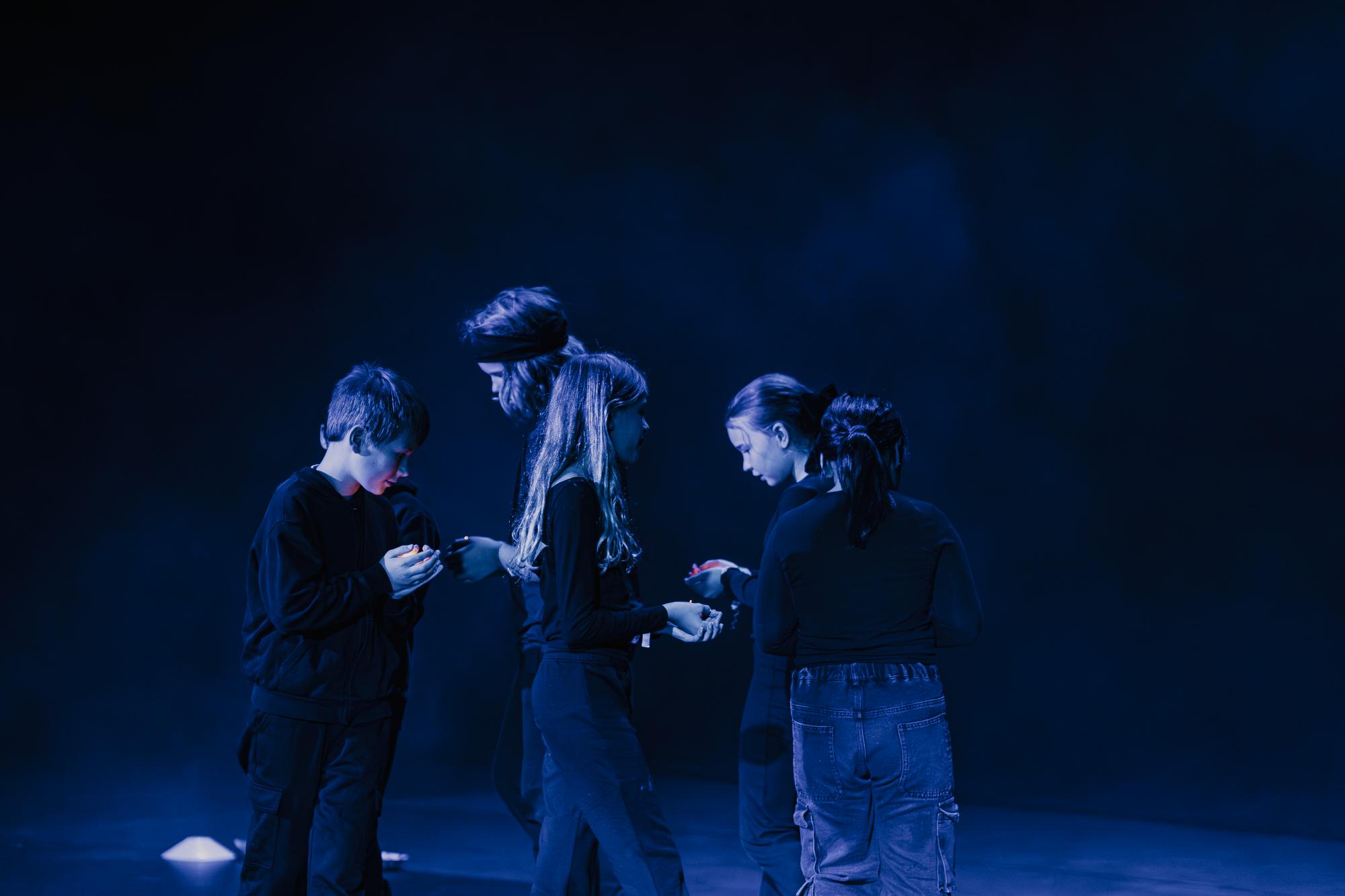 Fünf Schüler:innen, die in schwarzer Kleidung in einem engen Kreis gehen und blicken auf ihre Hände, aus denen ein Leuchten hervorgeht. Der Hintergrund ist dunkel.