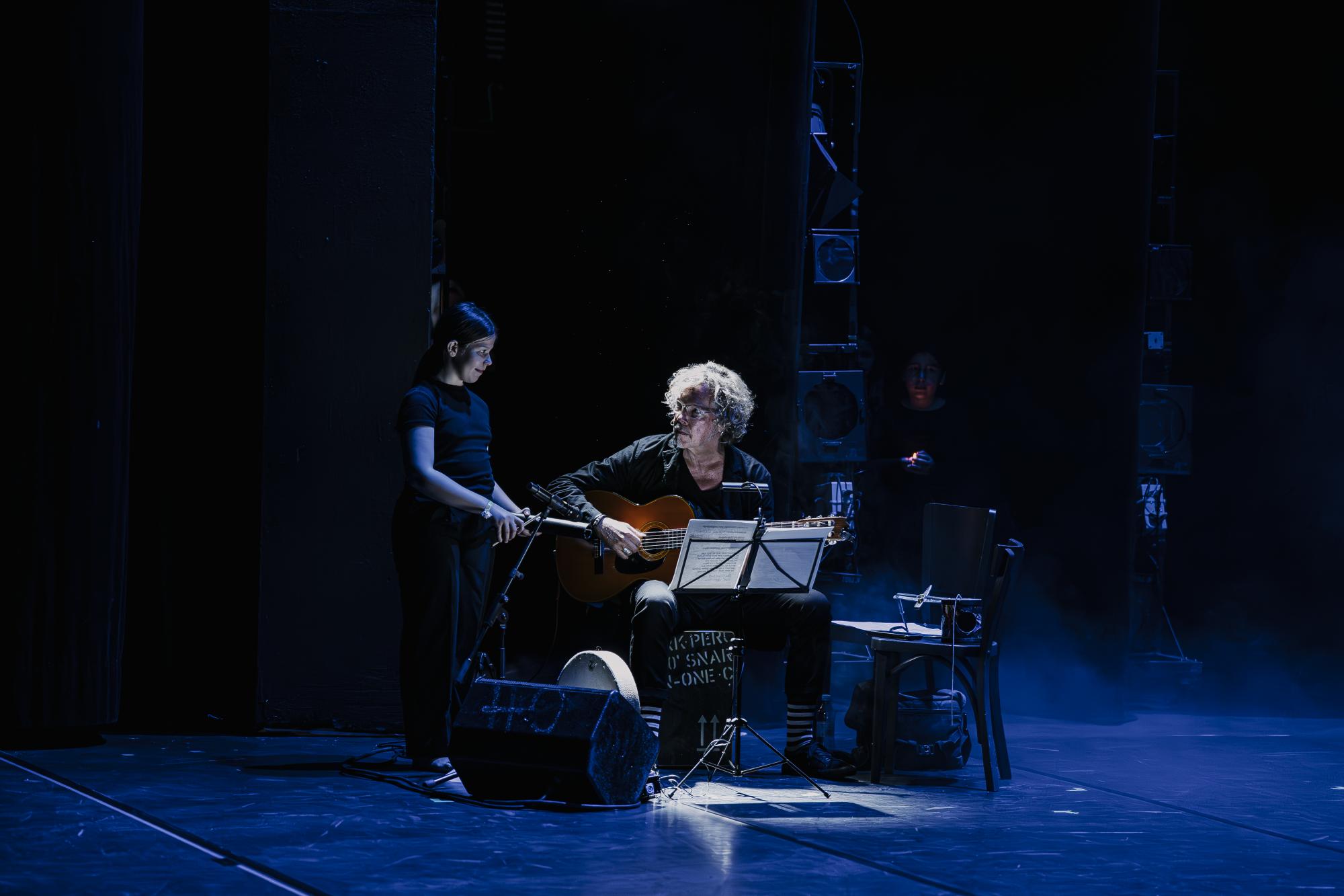 Zwei Menschen sind auf der Bühne. Ein Mann spielt Gitarre. Daneben steht ein Kind mit dem Instrument Kuhglocke. Die beiden schauen sich an.