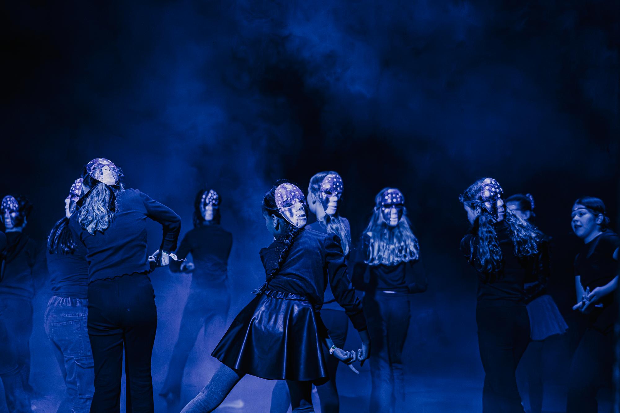 Zehn Schüler:innen stehen im blau ausgeleuchtetem Nebel auf der Bühne. An ihren Hinterköpfen haben sie Masken, auf die Fliegenpilze aufgemalt sind. In der Mitte des Bildes schauen eine Maske und ein Gesicht sich in direkt in die 