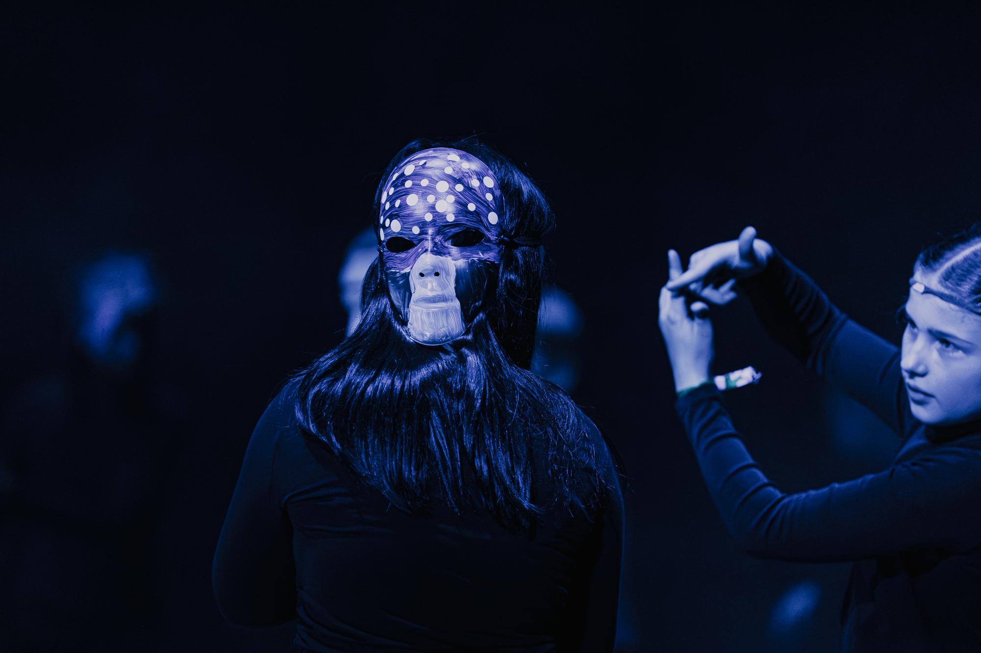 Zwei Personen: Eine überkreuzt ihre Finger zu dem Elfenzeichen und hält sie auf Augenhöhe der anderen Person, dessen Hinterkopf eine bemalte Fliegenpilzmaske ziert.