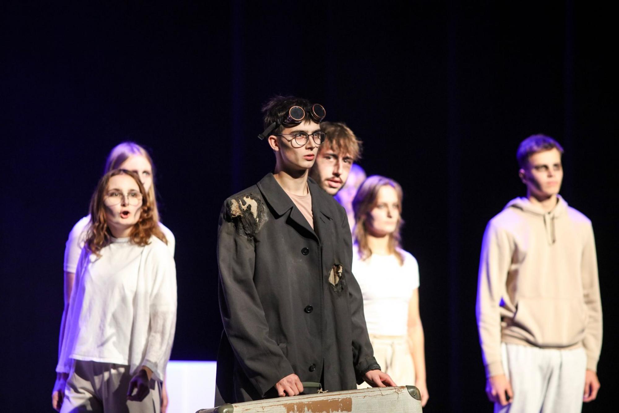 Im Vordergrund ist ein junger Mann in dunklem zerschlissenem Mantel, einer Steampunk Brille auf der Stirn mit einem Koffer in der Hand. Leicht verschwommen sieht man dahinter sechs Personen in heller Kleidung, die nach vorne blicken. Einer von ihnen schaut den Mann mit gerunzelter Stirn an.