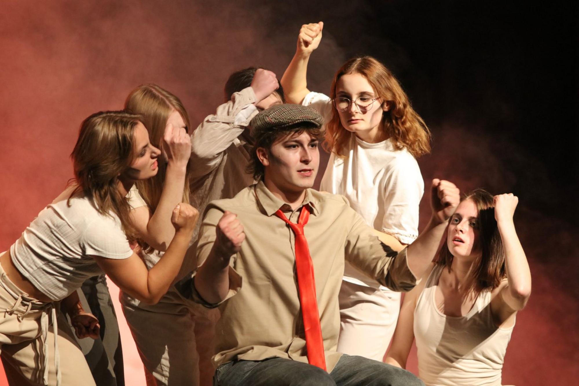 Vor orangenem Nebel sitzt ein junger Mann mit Schiebermütze und roter Krawatte. Neben und hinter ihm sind fünf junge Frauen, die jeweils eine Faust hochhalten und ihn angucken. Zwei von ihnen haben leicht geöffnete Münder.